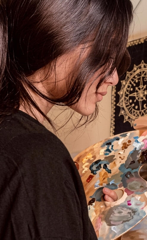 Maya Valasai Maya Valasai holding an artist palette and paintbrush, working on a canvas painting in her studio.